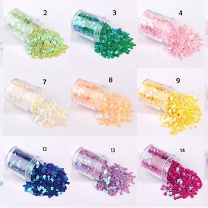 May include: A collection of fifteen small, clear plastic jars, each filled with colorful sequins. The sequins are in various colors, including green, pink, blue, yellow, orange, and purple. The jars are tilted, with sequins spilling out.