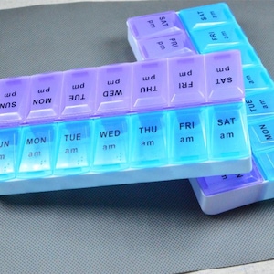 Pode incluir: Dois organizadores semanais de comprimidos em cores gradientes azul e roxo. Cada organizador tem sete compartimentos com os dias da semana e as indicações "am" e "pm". Os organizadores estão em uma superfície cinza.
