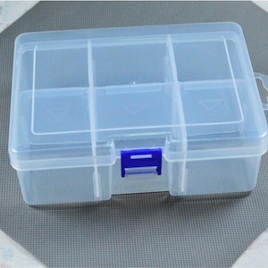 Puede incluir: Una caja de almacenamiento de plástico transparente con tapa con bisagras y seis compartimentos. La caja tiene un cierre azul y un asa pequeña. La caja está sobre una superficie gris.