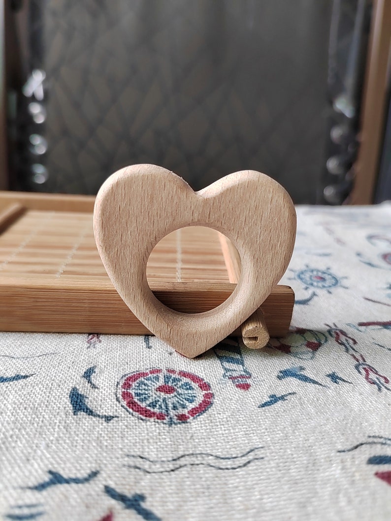 May include: A light brown wooden heart-shaped teether for babies. The teether has a smooth surface and a hole in the center.