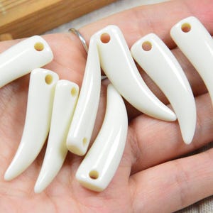 Op de afbeelding: Een verzameling ivoorkleurige, gebogen hangers die op hoektanden lijken. Elke hanger heeft een klein gaatje aan de bovenkant. De hangers worden op een hand getoond, wat suggereert dat ze worden gepresenteerd of te koop worden aangeboden.