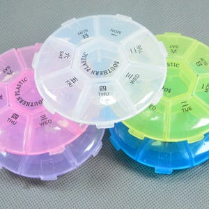 May include: Three multi-coloured, round pill organisers. One is pink and purple, another is clear, and the third is blue and green. Each has seven compartments labelled with days of the week and the word "PLASTIC".