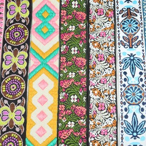 Könnte beinhalten: Sieben vertikale Streifen aus besticktem Stoffbesatz mit verschiedenen Mustern. Die Designs umfassen florale Motive in Pink, schwarze Bänder mit gelben und violetten Kreismustern, geometrische Formen in Gelb und Pink, grüne Blumenmuster sowie blaue botanische Designs.