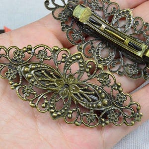 Könnte beinhalten: Zwei antike, bronzefarbene Haarspangen. Die Spangen weisen ein durchbrochenes Design mit Blumen- und Blättermustern auf. Die Metall-Haarspangen sind auf der Rückseite der Spangen sichtbar. Die Spangen sind etwa 7.6 cm lang.