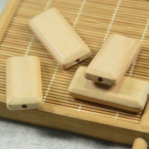 Puede incluir: Cuatro cuentas rectangulares de madera natural con un agujero perforado en el centro. Las cuentas están dispuestas sobre una estera de bambú.