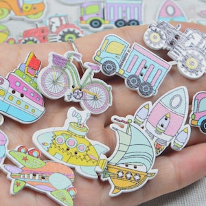 May include: A selection of colourful wooden buttons featuring transport designs such as a ship, bicycle, truck, rocket, and submarine. The buttons are painted in pastel shades, creating a whimsical and playful aesthetic, ideal for craft projects.