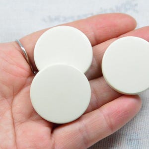 May include: Three off-white, circular buttons are shown. The buttons are smooth and opaque, with a slightly rounded edge. The buttons are a solid, neutral colour, and appear to be made of plastic or a similar material.