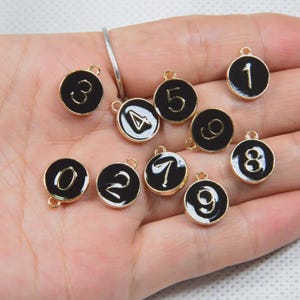 Könnte beinhalten: Eine Sammlung von schwarzen und goldenen Zahlenanhängern, jeweils mit einer anderen Zahl von 0 bis 9. Die Zahlen sind in weißer Schrift auf schwarzem Hintergrund und von einem goldenen Rand umgeben. Jeder Anhänger hat eine kleine Schlaufe zum Befestigen.
