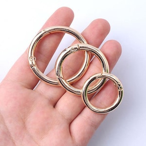 May include: Three gold-colored metal spring rings of varying sizes. The rings are round with a hinged opening and a secure clasp. The rings are shown in a hand, suggesting their size and use for attaching items.