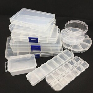 Puede incluir: Una colección de recipientes de almacenamiento de plástico transparente de varios tamaños. Varias cajas rectangulares con tapas con bisagras están apiladas. Otros recipientes incluyen una pequeña caja cuadrada, un recipiente redondo con múltiples compartimentos y varias cajas largas y estrechas con compartimentos numerados.