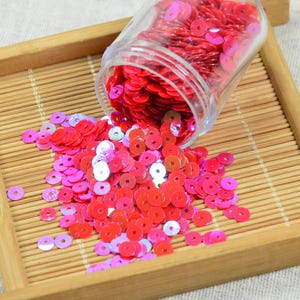 May include: A clear jar of red sequins is tipped over, spilling onto a bamboo tray. The sequins are a mix of red and pink, with some iridescent. The tray is made of light brown bamboo and sits on a neutral fabric.