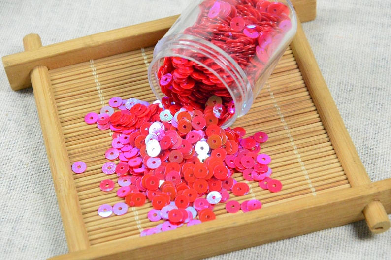 May include: A clear jar of red sequins is tipped over, spilling onto a bamboo tray. The sequins are a mix of red and pink, with some iridescent. The tray is made of light brown bamboo and sits on a neutral fabric.