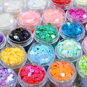 May include: A selection of small, clear plastic pots filled with colourful sequins. The sequins are in various shades, including pink, blue, green, yellow, and purple. The pots are arranged closely together, creating a vibrant display.