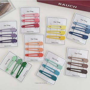Könnte beinhalten: Eine Auswahl an farbigen Haarspangen auf weißen Karten. Jede Karte zeigt drei Spangen in den Farbtönen Rosa, Rot, Gelb, Blau, Grün, Orange und Braun. Die Karten sind mit "Xue Ning" und "Made in China" beschriftet.
