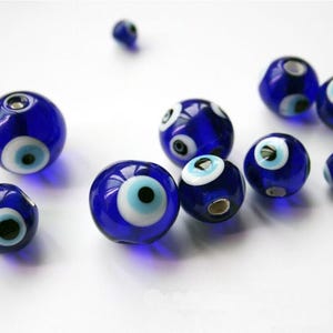 Könnte beinhalten: Eine Ansammlung blauer Glasperlen mit einem weißen und hellblauen Augendesign. Die Perlen sind rund und haben eine glänzende Oberfläche. Die Perlen sind auf einer weißen Oberfläche verstreut.