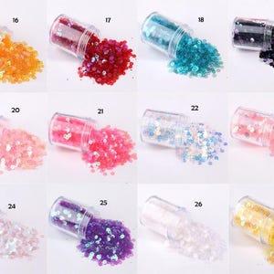 May include: A collection of small, clear jars, each filled with colorful sequins. The sequins are in various colors, including orange, red, teal, black, pink, light blue, and purple. The jars are tipped over, spilling the sequins.