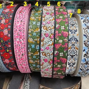 Könnte beinhalten: Eine Sammlung von neun Rollen dekorativem Webband, die jeweils unterschiedliche Muster wie geometrische Formen, florale Motive und komplizierte Schnörkel in verschiedenen Farben wie Rot, Rosa, Grün, Blau und Weiß aufweisen und nebeneinander angeordnet sind.