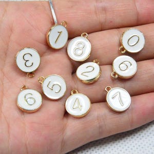 Könnte beinhalten: Eine Sammlung von goldgerahmten, weißen Emaille-Zahlenanhängern, jeder mit einer anderen Ziffer von Null bis Acht. Die Zahlen sind in goldener Schrift. Jeder Anhänger hat eine kleine Schlaufe zum Befestigen an einer Kette oder einem Armband.