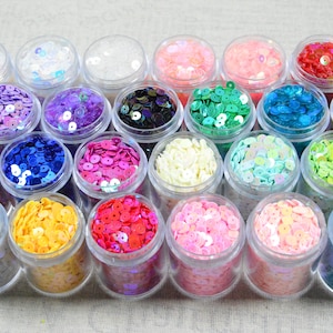 May include: A collection of small, clear plastic jars filled with colorful sequins. The sequins are in various colors, including pink, blue, green, yellow, and purple. The jars are arranged closely together, creating a vibrant display.