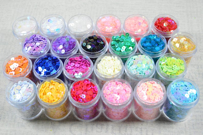 May include: A collection of small, clear plastic jars filled with colorful sequins. The sequins are in various colors, including pink, blue, green, yellow, and purple. The jars are arranged closely together, creating a vibrant display.