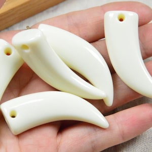 May include: Six off-white, curved pendants, each shaped like a fang. They have a smooth, glossy surface and a small hole at the top for threading. The pendants are shown against a neutral backdrop.