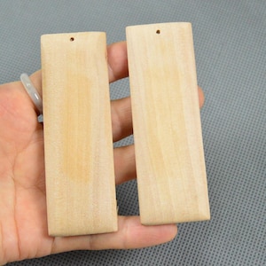 May include: Two rectangular wooden pendants with a light, natural wood grain. Each pendant has a small hole at the top for stringing. The pendants are held in a hand against a gray background.