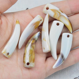 May include: A collection of seven curved, tooth-shaped pendants made of smooth, polished material. Each piece features a glossy finish with a marbled pattern in shades of cream, white, and golden brown, and includes a small circular hole at the top.