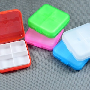 Puede incluir: Una colección de pastilleros cuadrados y coloridos. Uno está abierto, revelando cuatro compartimentos. Los otros pastilleros están cerrados, en tonos de rojo, verde, rosa, azul y blanco. Estos pequeños recipientes de plástico están diseñados para organizar medicamentos.