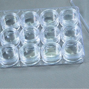 May include: A clear plastic container with twelve small, round, empty jars arranged in a grid. The jars have screw-on lids and are designed for storing small items like cosmetics or craft supplies. The container is transparent.