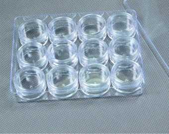 May include: A clear plastic container with twelve small, round, empty jars arranged in a grid. The jars have screw-on lids and are designed for storing small items like cosmetics or craft supplies. The container is transparent.