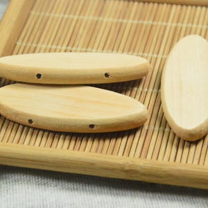 Könnte beinhalten: Drei ovale Holzperlen mit jeweils zwei Löchern. Die Perlen sind hellbraun und liegen auf einer Bambusmatte.
