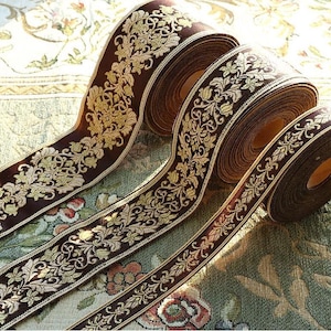 Könnte beinhalten: Nahaufnahme von mehreren Rollen dekorativen Bandes. Das Band ist tiefbraun mit einem verzierten goldenen Blumenmuster. Die Kanten sind mit einer dünnen goldenen Bordüre versehen. Die Rollen sind diagonal angeordnet und zeigen das detaillierte Design.