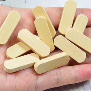 May include: A handful of small, rectangular wooden beads with rounded ends. The beads are a light, natural wood colour and appear to be unfinished. They are held in a hand, suggesting they are craft supplies or components for jewellery making.