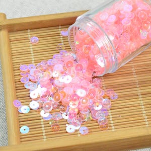 May include: A clear plastic jar tipped over, spilling iridescent pink sequins onto a small, light brown bamboo tray. The sequins are round and vary in size, reflecting light with a subtle shimmer. The tray has a rectangular shape.