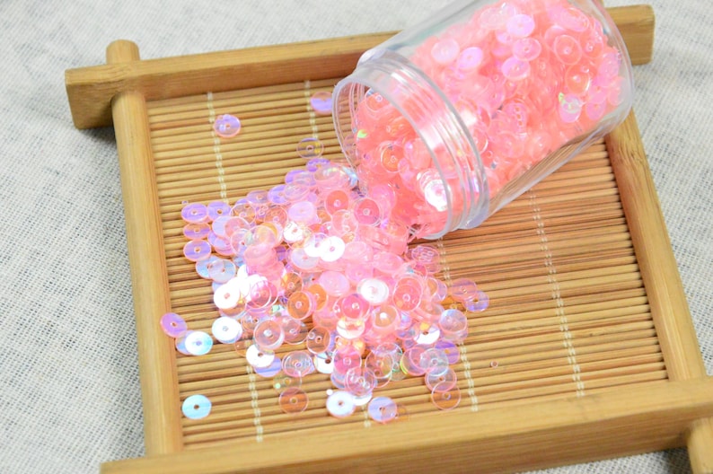 May include: A clear plastic jar tipped over, spilling iridescent pink sequins onto a small, light brown bamboo tray. The sequins are round and vary in size, reflecting light with a subtle shimmer. The tray has a rectangular shape.