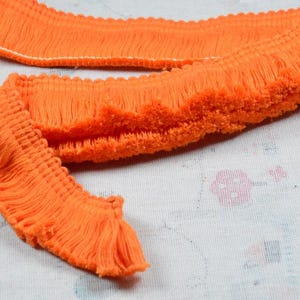 Puede incluir: Primer plano de un ribete de flecos naranja brillante. El ribete tiene un borde texturizado y en bucle y una franja de hilos sueltos y suaves. El ribete está sobre una tela blanca estampada.