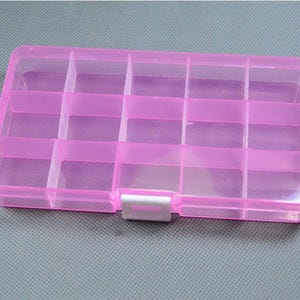 May include: A translucent pink plastic organizer box with multiple compartments. The box has a rectangular shape with a hinged lid and a white clasp. Ideal for storing small items like beads or jewelry.