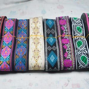 Könnte beinhalten: Eine Sammlung von acht Rollen dekorativem Stoffband, die nebeneinander angeordnet sind. Jede Rolle weist unterschiedliche gestickte Muster auf, darunter florale, geometrische und Paisley-Motive in leuchtenden Farben wie Pink, Blau, Grün, Gold und Rot.