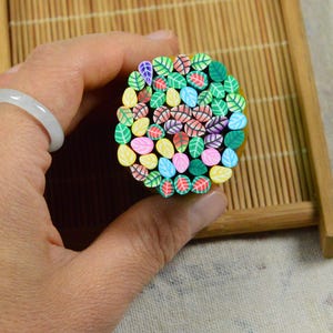 May include: A hand holds a circular arrangement of colorful polymer clay leaves. The leaves are various colors, including green, red, pink, yellow, and blue. A white ring is on the finger. The arrangement is on a bamboo tray.