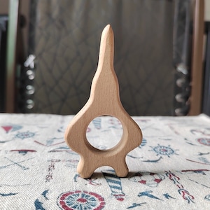 May include: A wooden teether shaped like a rocket ship with a ring in the middle. The teether is light brown and has a smooth finish.