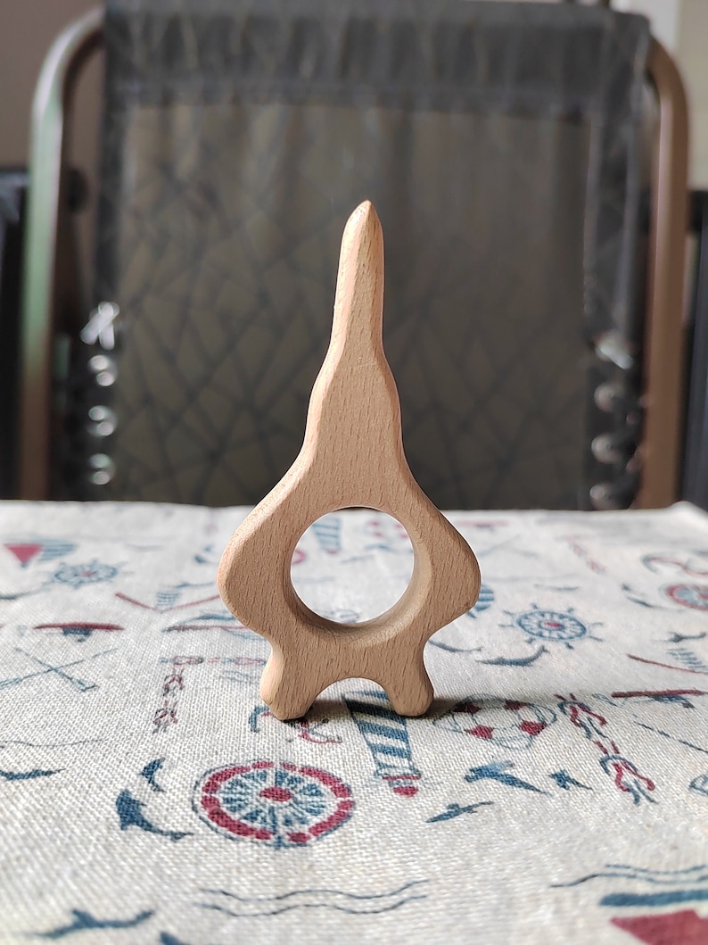 May include: A wooden teether shaped like a rocket ship with a ring in the middle. The teether is light brown and has a smooth finish.