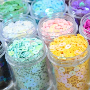 May include: A selection of small, clear jars filled with colourful sequins. The sequins are in various colours, including green, blue, pink, yellow, and purple. The sequins are round and have a hole in the centre. Suitable for crafts and embellishments.