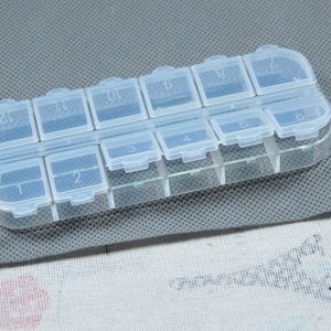 Peut inclure: Un pilulier en plastique transparent avec douze compartiments num&eacute;rot&eacute;s. Chaque compartiment a un couvercle &agrave; charni&egrave;re. L'organisateur est rectangulaire et repose sur une surface grise.