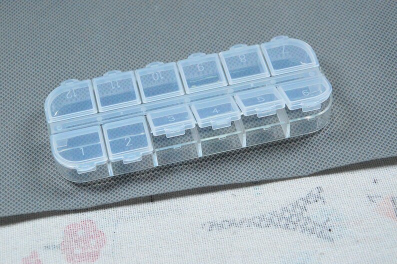 Peut inclure: Un pilulier en plastique transparent avec douze compartiments num&eacute;rot&eacute;s. Chaque compartiment a un couvercle &agrave; charni&egrave;re. L'organisateur est rectangulaire et repose sur une surface grise.