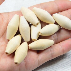 May include: A collection of ten light beige, unfinished wooden beads with a smooth, elongated, spindle-like shape. Each bead features a small, centered hole running through its length, showcasing a natural, matte texture and pale wood grain.