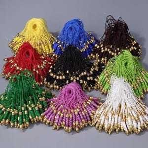 May include: A collection of colorful tassels with gold-colored beads. The tassels are in various colors, including yellow, red, blue, black, brown, green, pink, and white. Each tassel has a loop at the top and a beaded bottom.