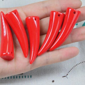 Op de afbeelding: Vijf gladde, glanzende, felrode hoornvormige hangers, elk voorzien van een klein rond gaatje aan de bredere kant om aan een koord te rijgen. Deze gebogen, taps toelopende onderdelen hebben een gepolijste afwerking en een egale rode kleur.