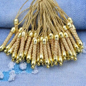 May include: A cluster of gold-coloured tassels, each with a braided cord and metallic cap. The tassels are bunched together, with the cords spreading out. The background is a light blue denim fabric.
