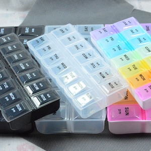 May include: A collection of weekly pill organisers in various colours. Each organiser is rectangular with compartments labelled with days of the week and AM/PM. Colours include black, clear, and pastel shades.