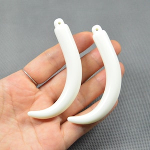 May include: Two smooth, curved, off-white pendants shaped like animal tusks or teeth. Each piece features a small, circular hole at the top end for attachment, with a polished, solid surface and a tapered, pointed tip at the bottom end.